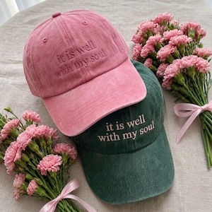 May include: Two baseball caps, one pink and one dark green, are displayed. The pink cap has the embroidered text "it is well with my soul." The green cap also has the same text. Pink carnations and a ribbon are in the background.