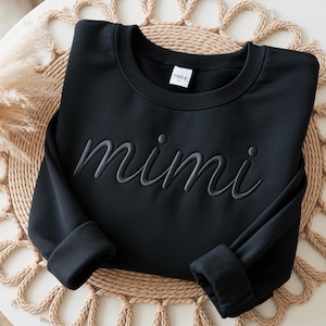 May include: A black long-sleeve sweatshirt featuring the word mimi embroidered in a matching black thread across the chest. The garment has a crew neckline, a white interior brand label, and long sleeves with folded cuffs at the ends.