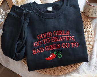 Może przedstawiać: Czarna bluza z okrągłym dekoltem i czerwonym haftowanym napisem GOOD GIRLS GO TO HEAVEN, BAD GIRLS GO TO, po którym następuje wyhaftowana czerwona papryczka chili i zielona litera S. Bluza ma długie rękawy i prążkowany kołnierzyk.