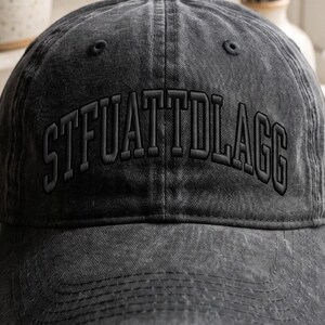 K&ouml;nnte beinhalten: Eine dunkelgraue, ausgewaschene Baseballkappe mit dem gestickten Text "STFUATTDLAGG" in passendem Dunkelgrau. Die Kappe hat einen gebogenen Schirm und ein klassisches Sechs-Panel-Design.
