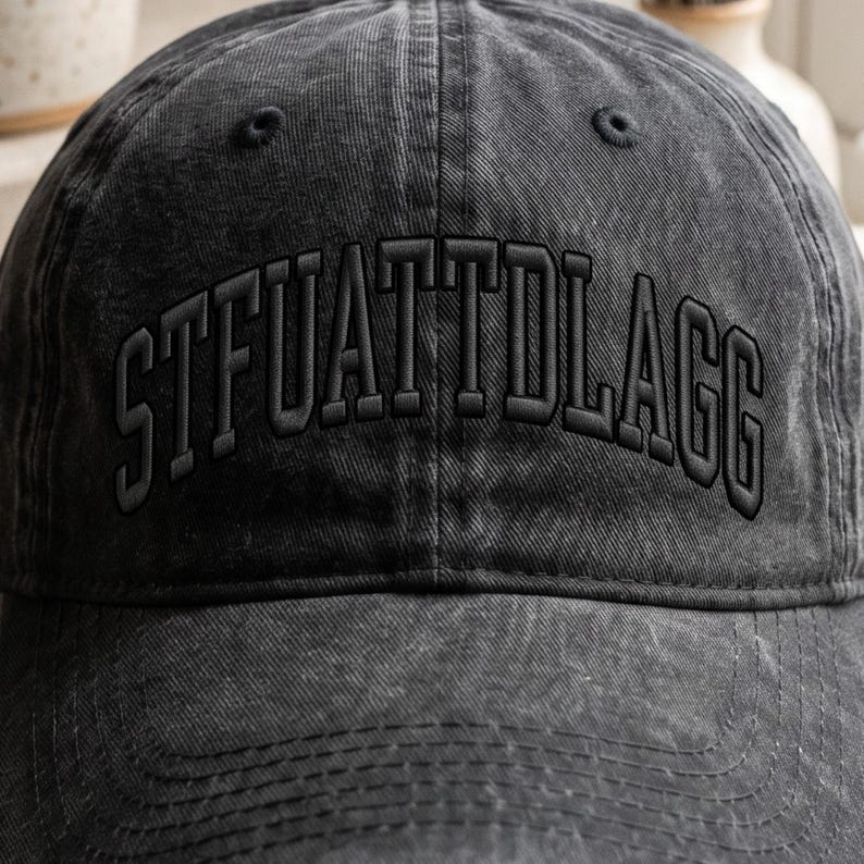 K&ouml;nnte beinhalten: Eine dunkelgraue, ausgewaschene Baseballkappe mit dem gestickten Text "STFUATTDLAGG" in passendem Dunkelgrau. Die Kappe hat einen gebogenen Schirm und ein klassisches Sechs-Panel-Design.
