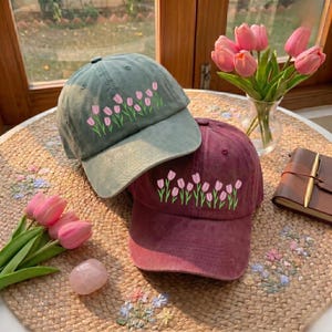 May include: Two baseball caps, one sage green and one burgundy, are displayed. Both caps feature an embroidered design of pink tulips. The caps are on a woven mat, with a vase of pink tulips and a small notebook nearby.