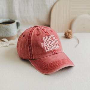 May include: A red baseball cap with the text "GOD'S FAVORITE LESBIAN" in white block letters. The cap has a distressed look with visible stitching and a curved brim. The hat is made of a soft, worn material.