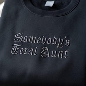 May include: A dark navy blue sweatshirt with the embroidered text "Somebody's Feral Aunt" in a gothic font. The crew neck sweatshirt is made of a soft material.