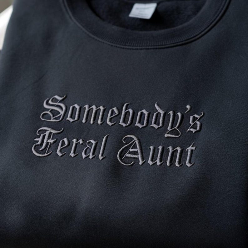 May include: A dark navy blue sweatshirt with the embroidered text "Somebody's Feral Aunt" in a gothic font. The crew neck sweatshirt is made of a soft material.