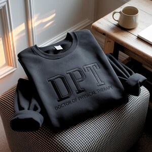 May include: A black sweatshirt with the letters "DPT" embroidered on the front, along with the words "DOCTOR OF PHYSICAL THERAPY". The sweatshirt is folded on a patterned ottoman. A mug sits on a wooden table in the background.