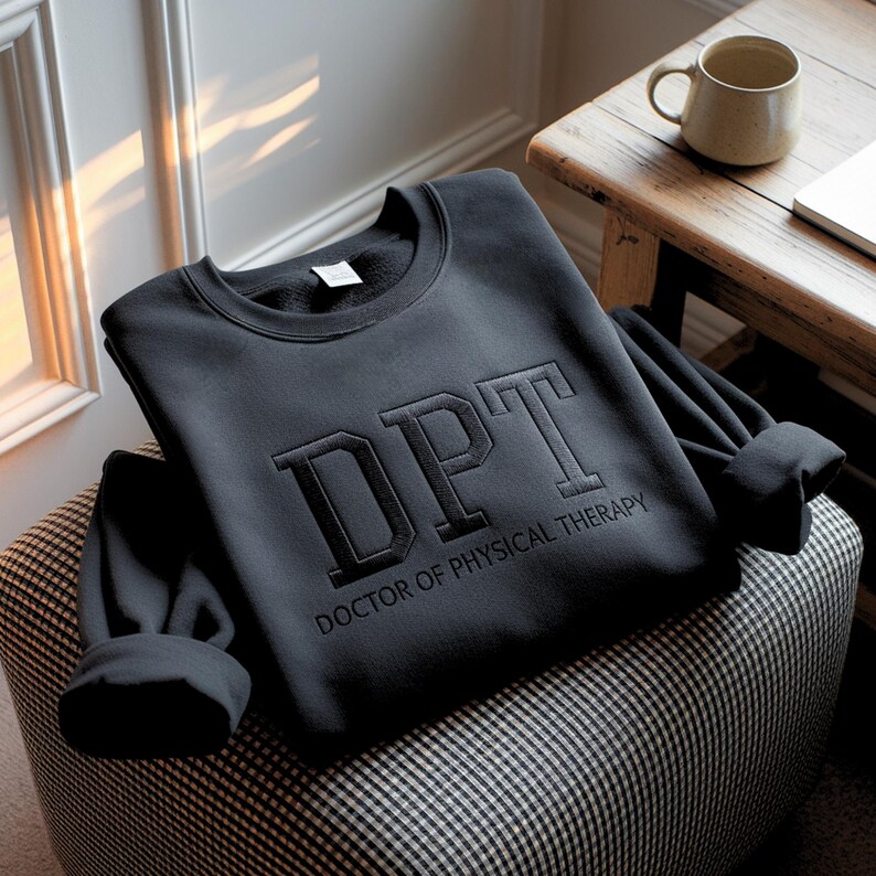 May include: A black sweatshirt with the letters "DPT" embroidered on the front, along with the words "DOCTOR OF PHYSICAL THERAPY". The sweatshirt is folded on a patterned ottoman. A mug sits on a wooden table in the background.