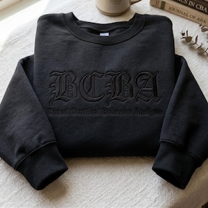 May include: A black crewneck sweatshirt featuring the embroidered text BCBA in a large gothic font, with the words Board Certified Behavior Analyst embroidered in a smaller, standard sans-serif font directly underneath the larger letters.
