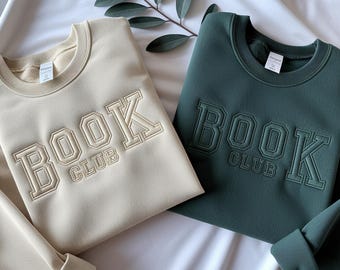 May include: Two crewneck sweatshirts, one in beige and one in dark green, each featuring the embroidered text "BOOK CLUB" in a collegiate font across the chest. Both garments have long sleeves and ribbed necklines, cuffs, and hems.
