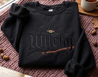 Może przedstawiać: Czarna bluza z okrągłym dekoltem i haftowanym wzorem z przodu. Haft przedstawia słowo Witchy stylizowaną czcionką, kapelusz czarownicy ze złotą klamrą, miotłę oraz małe motywy nietoperzy i gwiazd wykonane czarną nicią.