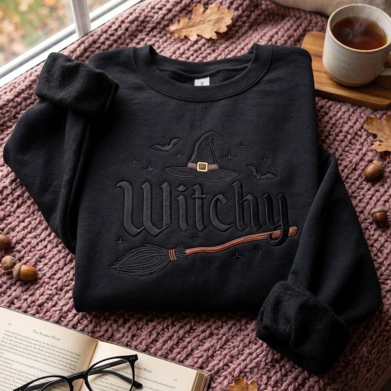 May include: A black crewneck sweatshirt featuring an embroidered design on the front. The embroidery includes the word Witchy in a stylized font, a witch hat with a gold buckle, a broomstick, and small bat and star motifs in black thread.