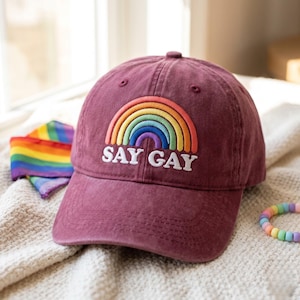 May include: A maroon baseball cap featuring an embroidered rainbow graphic above the white text SAY GAY. The cap has a structured crown and a curved brim, with a small rainbow-coloured fabric piece and a pastel beaded bracelet nearby.