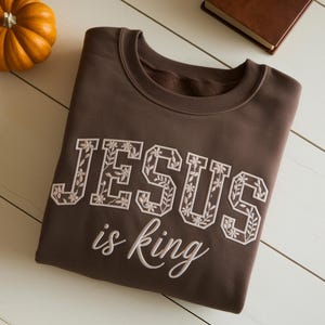 May include: A brown sweatshirt with the words "JESUS is king" embroidered in white. The letters are decorated with floral designs. A small orange pumpkin is in the upper left corner.