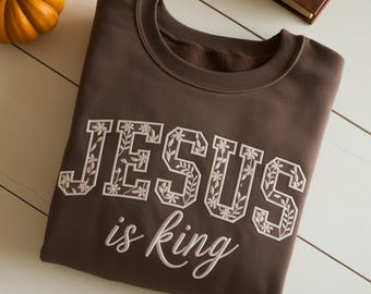 May include: A brown sweatshirt with the words "JESUS is king" embroidered in white. The letters are decorated with floral designs. A small orange pumpkin is in the upper left corner.