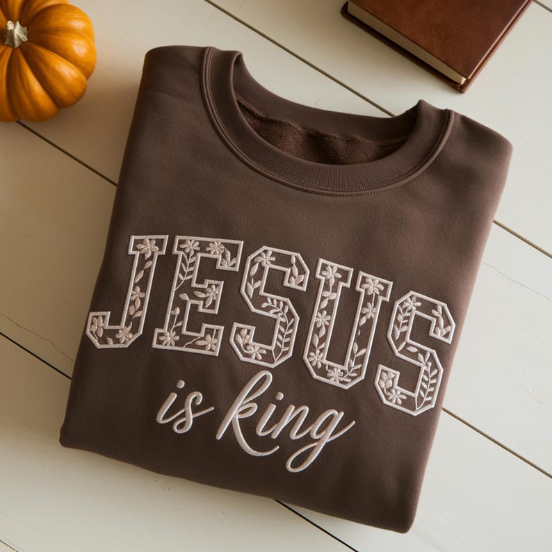May include: A brown sweatshirt with the words "JESUS is king" embroidered in white. The letters are decorated with floral designs. A small orange pumpkin is in the upper left corner.