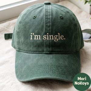 May include: A dark green baseball cap with a six-panel construction and a curved brim. The front features the embroidered text "i'm single." in a light beige, sans-serif font. The cap has a fabric strap with a metal buckle on the back.