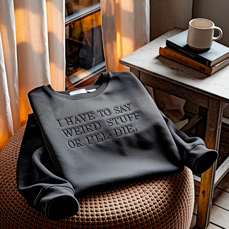 Puede incluir: Una sudadera gris oscuro con el texto bordado "I HAVE TO SAY WEIRD STUFF OR I'LL DIE". La sudadera est&aacute; doblada sobre un otom&aacute;n tejido. Una peque&ntilde;a mesa de madera sostiene una taza y dos libros.