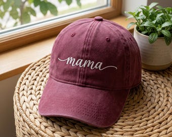 Puede incluir: Gorra de béisbol burdeos con la palabra "mama" bordada en escritura blanca. La gorra está colocada sobre una cesta tejida, con una pequeña planta en maceta en el fondo. La gorra tiene una visera curva y un botón en la parte superior.
