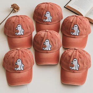 May include: Six terracotta-colored baseball caps with a ghost and pumpkin patch design. The caps are arranged in a circular pattern, with the ghost holding a pumpkin. The caps have a curved brim and a classic six-panel design.