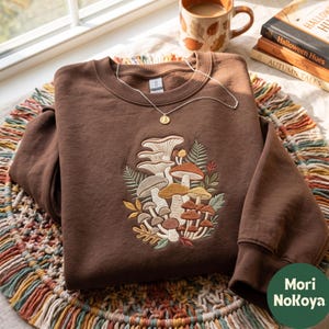 Könnte beinhalten: Ein dunkelbraunes Sweatshirt mit Rundhalsausschnitt und einer Stickerei aus verschiedenen Pilzen und Farnen in Beige-, Orange- und Grüntönen. Eine zarte goldfarbene Halskette mit einem kleinen runden Anhänger liegt am Halsausschnitt des Kleidungsstücks.