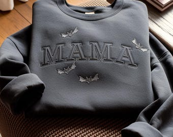 May include: Dark grey sweatshirt featuring the embroidered word "MAMA" in a collegiate font, with bat motifs. The sweatshirt is folded on a patterned surface, with a wooden table and a book in the background.