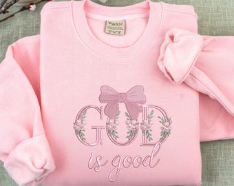 Op de afbeelding: Een lichtroze sweatshirt met de geborduurde zin "GOD is good". Het woord "GOD" is versierd met een roze strik en bloemmotieven. De top met lange mouwen is netjes opgevouwen.