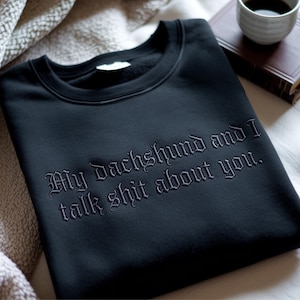 May include: Black sweatshirt with the embroidered text "My dachshund and I talk shit about you." The sweatshirt is black and has a crew neck. A cup of coffee and a book are in the background.