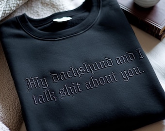 Może przedstawiać: Czarna bluza z okrągłym dekoltem i ciemnoszarym haftowanym napisem o treści: My dachshund and I talk shit about you. Odzież posiada prążkowany kołnierz i długie rękawy, a haft znajduje się na środku klatki piersiowej materiału.