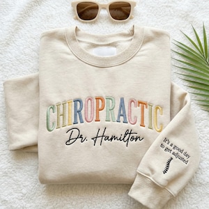 May include: A beige crewneck sweatshirt featuring the word CHIROPRACTIC embroidered in multicolored pastel letters, with the text Dr. Hamilton and the phrase It's a good day to get adjusted with a spinal graphic on the sleeve.