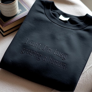 May include: A black crewneck sweatshirt featuring tonal black embroidery on the chest. The text reads, "I can't, I'm busy growing a human." The garment has a ribbed collar and long sleeves, showcasing a clean, monochromatic design with bold lettering.