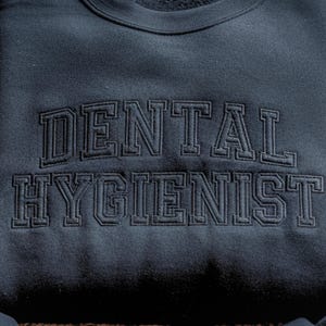 Peut inclure: Sweat-shirt bleu marine fonc&eacute; avec l'inscription "DENTAL HYGIENIST" brod&eacute;e en fil bleu marine fonc&eacute; assorti. Le texte est &eacute;crit dans une police de style universitaire.