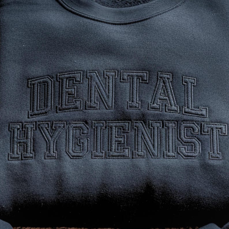 Peut inclure: Sweat-shirt bleu marine fonc&eacute; avec l'inscription "DENTAL HYGIENIST" brod&eacute;e en fil bleu marine fonc&eacute; assorti. Le texte est &eacute;crit dans une police de style universitaire.