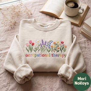 May include: A beige crewneck sweatshirt featuring an embroidered design of colorful wildflowers and the text "occupational therapy" in multi-colored lowercase letters. The embroidery includes tulips, daisies, lavender, and various small blossoms.
