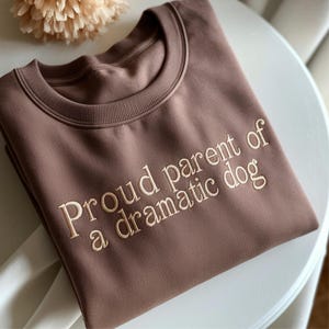 May include: A brown sweatshirt with the embroidered text "Proud parent of a dramatic dog." The crew neck sweatshirt is made of a soft material and is a neutral color. The text is in a light beige color.