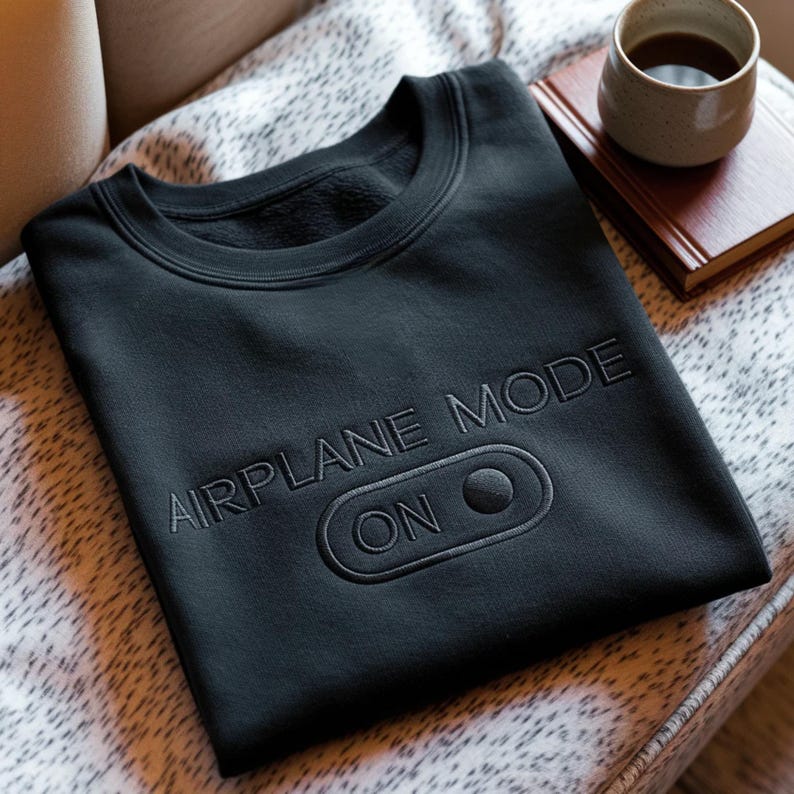 Puede incluir: Sudadera negra de cuello redondo con la inscripci&oacute;n bordada "AIRPLANE MODE ON" y un s&iacute;mbolo de interruptor de encendido/apagado. El dise&ntilde;o est&aacute; realizado con un hilo oscuro a juego. La sudadera se muestra sobre una superficie estampada.