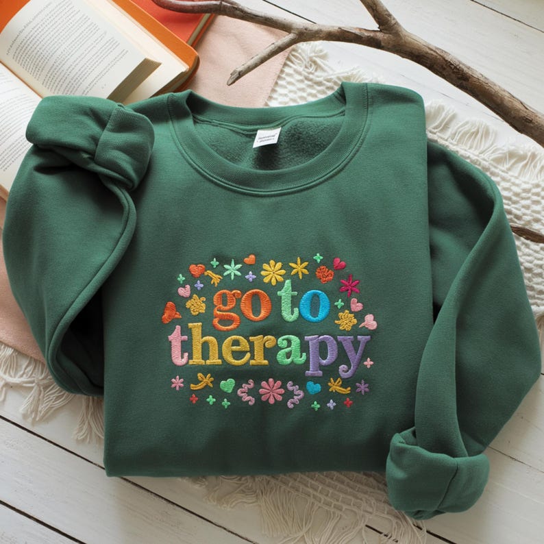 May include: A dark green sweatshirt with the embroidered phrase "go to therapy" in colorful letters, surrounded by floral and heart designs. The long-sleeved top is folded neatly, showcasing the design.