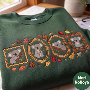 Könnte beinhalten: Ein dunkelgrünes Sweatshirt mit Rundhalsausschnitt, das mit Koalas in vier verschiedenen dekorativen Rahmen bestickt ist. Das Design enthält verstreute Herbstblätter in Rot-, Orange- und Gelbtönen. Der Text Mori NoKoya ist in einem Kreis zu sehen.