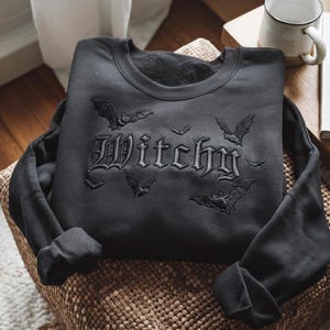 Könnte beinhalten: Ein schwarzes Sweatshirt mit Rundhalsausschnitt und einer schwarzen Stickerei des Wortes Witchy in gotischer Schrift, umgeben von mehreren gestickten Fledermäusen unterschiedlicher Größe, die um den Text fliegen. Das Sweatshirt hat lange Ärmel.