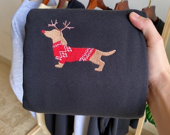 May include: A folded black sweatshirt featuring an embroidered dachshund wearing a red sweater with a white pattern and antlers. The dog is brown with a red nose. Other sweatshirts hang in the background.