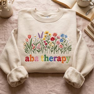 May include: A beige crewneck sweatshirt featuring an embroidered design of colourful wildflowers in shades of pink, yellow, red, blue, and white. Below the floral arrangement, the text "aba therapy" is embroidered in multicoloured letters.