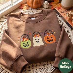 May include: A brown crewneck sweatshirt featuring an embroidered design of three Halloween-themed buckets filled with candy. The design includes a green jack-o'-lantern bucket, a white ghost-shaped bucket, and an orange jack-o'-lantern bucket.