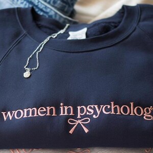 May include: A navy blue crewneck sweatshirt featuring the embroidered text "women in psychology" in a light pink thread, accompanied by a small embroidered pink bow icon centered directly below the text. A thin silver chain necklace rests on top.
