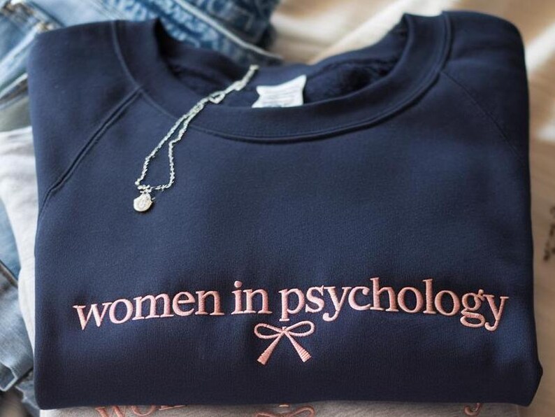 May include: A navy blue crewneck sweatshirt featuring the embroidered text "women in psychology" in a light pink thread, accompanied by a small embroidered pink bow icon centered directly below the text. A thin silver chain necklace rests on top.