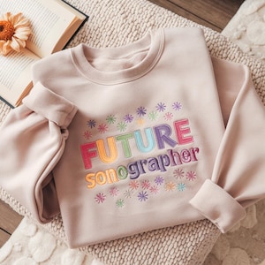 May include: A light pink sweatshirt with the embroidered text "FUTURE sonographer" in rainbow colors. The design includes colorful embroidered snowflakes. The sweatshirt is folded on a textured surface.