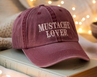Puede incluir: Gorra de béisbol granate con el texto "MUSTACHE LOVER" bordado en blanco. La gorra está sobre un libro. El fondo tiene una iluminación suave.