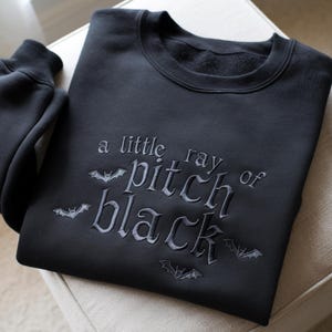 Op de afbeelding: Zwart sweatshirt met de geborduurde tekst "a little ray of pitch black" en vleermuismotieven. De donkergekleurde crewneck sweatshirt heeft een zacht, comfortabel uiterlijk.