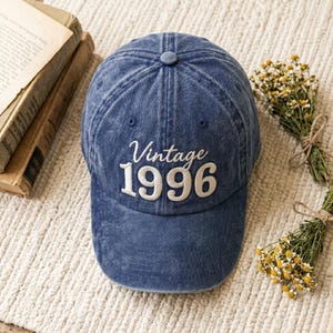 May include: A blue denim baseball cap with the text "Vintage 1996" embroidered in white. The cap has a curved brim and a button on top. The background includes books and dried flowers.