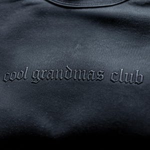 May include: A dark navy blue sweatshirt with the embroidered text "cool grandmas club" in a gothic font. The text is the same colour as the sweatshirt. The sweatshirt has a crew neck.