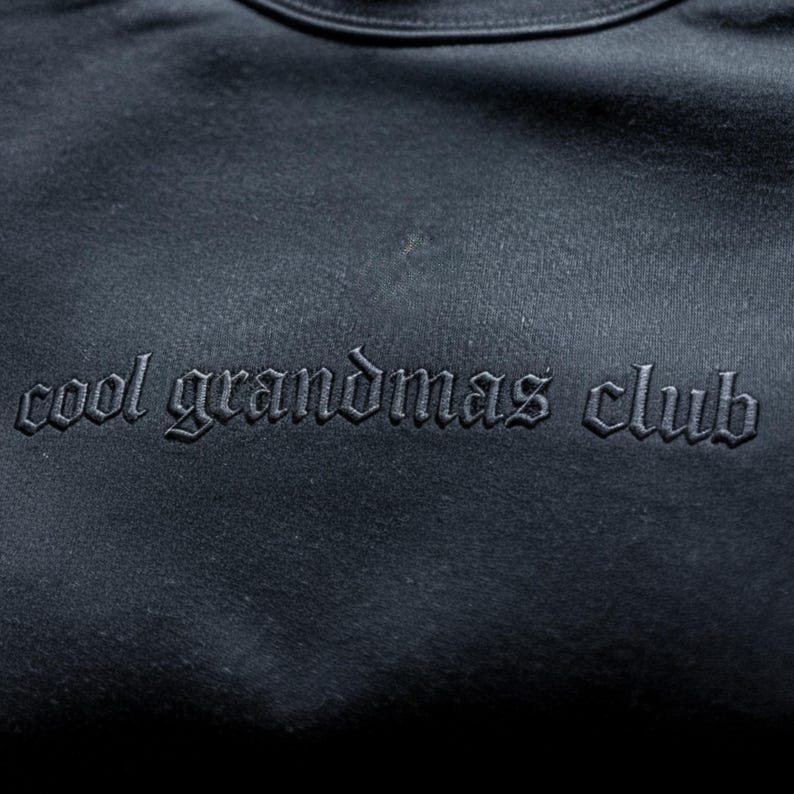May include: A dark navy blue sweatshirt with the embroidered text "cool grandmas club" in a gothic font. The text is the same colour as the sweatshirt. The sweatshirt has a crew neck.