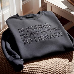 May include: Dark gray sweatshirt with the embroidered text "IF FOUND PLEASE RETURN TO LIBRARY". The crew neck sweatshirt is folded on a woven, round ottoman. The text is in a darker shade of gray.